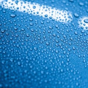Close-up of water droplets demonstrating the hydrophobic effect on a car surface after applying a ceramic coating. The image shows a glossy, well-protected blue car surface in a detailing studio