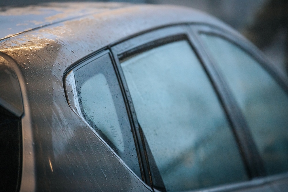 Side view of a car window covered with condensation and water droplets, possibly after rain or washing. The blurred background creates a soft, moody effect with plenty of copy space.